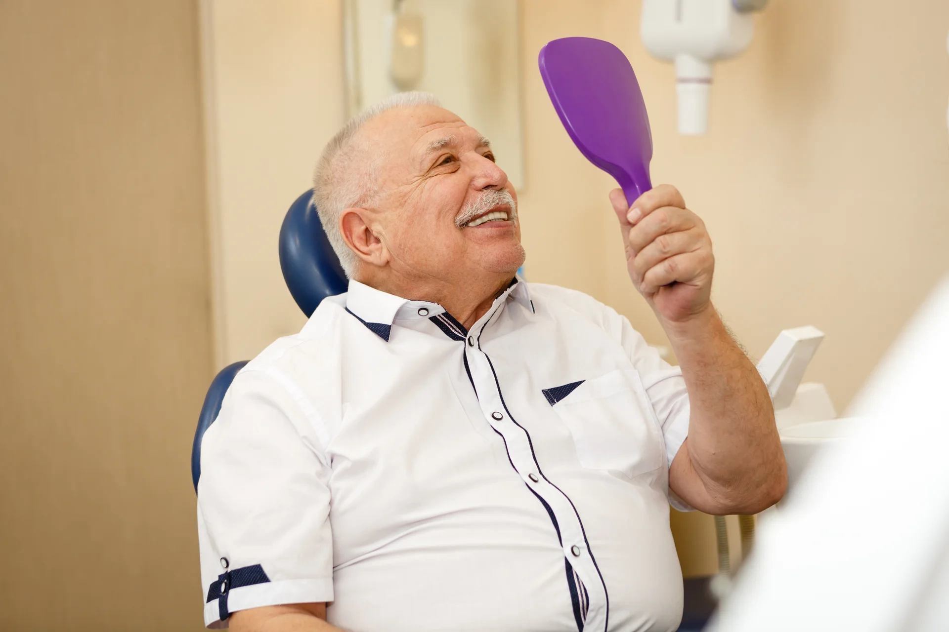 Older patient in the dental chair smiling at his new dentures in a hand mirror